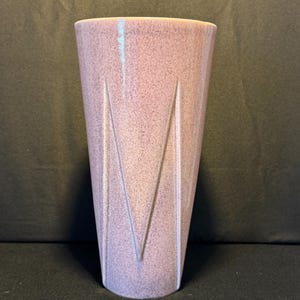 Puede incluir: Un jarrón de cerámica alto y cónico con un esmalte moteado de color rosa polvoriento. La superficie exterior presenta tres hendiduras verticales distintivas en forma de V que crean una apariencia geométrica y facetada en el cuerpo.
