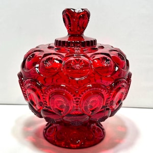 Könnte beinhalten: Eine leuchtend rote Glasschale für Süßigkeiten mit einem sich wiederholenden Mond- und Sternmuster auf dem abgerundeten Körper und Deckel. Das Stück enthält einen passenden Deckel mit einem dekorativen Knauf und steht auf einem runden Sockel.
