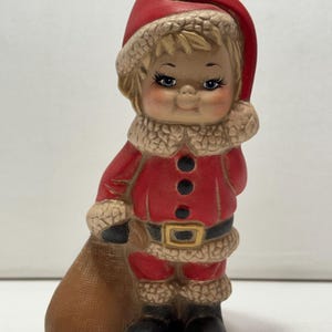 Könnte beinhalten: Eine Keramikfigur eines Kindes in einem roten Weihnachtsmannkostüm mit weißem, strukturiertem Besatz, passender roter Mütze und schwarzen Stiefeln. Die Figur hat blondes Haar, blaue Augen und hält einen großen braunen Sack an der Seite.