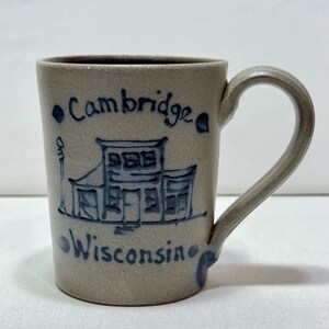 May include: A tan ceramic mug with a dark blue hand-painted illustration of a building and a street lamp. The text "Cambridge" is written above the building and "Wisconsin" is written below it in a dark blue script on the front surface.