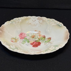May include: Oval porcelain dish with a floral pattern. The dish has a scalloped edge and is decorated with pink, red, and white roses and green leaves. The background has a gradient of cream, brown, and green.