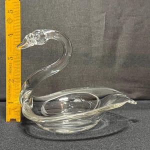 May include: Clear glass swan figurine. The swan has a long, curved neck and a bowl-shaped body. The figurine is approximately 4 inches long, as measured by a yellow ruler in the image. The swan is a decorative item.