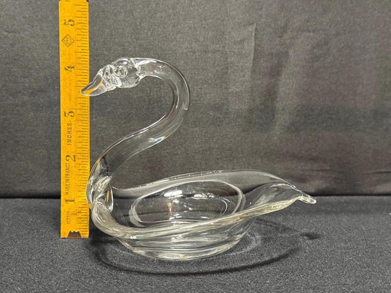May include: Clear glass swan figurine. The swan has a long, curved neck and a bowl-shaped body. The figurine is approximately 4 inches long, as measured by a yellow ruler in the image. The swan is a decorative item.