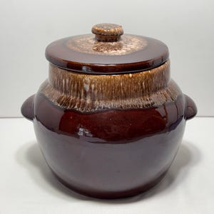 Może przedstawiać: Okrągły, ciemnobrązowy garnek ceramiczny z pasującą pokrywką i dwoma bocznymi uchwytami. Górna część korpusu oraz pokrywka posiadają dekoracyjny efekt ściekającego szkliwa w jaśniejszych odcieniach beżu i kremu, kontrastujący z ciemną podstawą.
