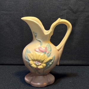 May include: A vintage, cream-colored pitcher with a floral design. The pitcher features a yellow flower with pink and green accents. It has a handle and a spout, and sits on a scalloped base. The pitcher is likely made of ceramic.