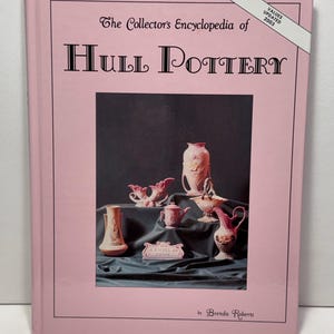 Puede incluir: Un libro de tapa dura titulado The Collector's Encyclopedia of Hull Pottery de Brenda Roberts. La cubierta rosa claro presenta una fotografía central de varios jarrones y jarras de cerámica rosa y blanca, con una banda blanca que dice Values Updated 2003.