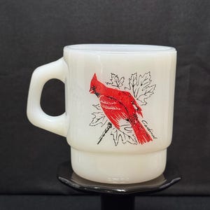 Puede incluir: Taza de cerámica blanca con un diseño de pájaro cardenal rojo y detalles de hojas negras. La palabra "Cardinal" está impresa en la taza. La taza tiene un asa y un diseño apilado. La taza está sobre un soporte negro.