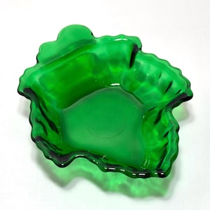 May include: A vibrant green glass dish with a unique, organic shape. The dish has a ruffled edge and a glossy finish, creating a striking visual appeal. The design features a central well and a textured surface.