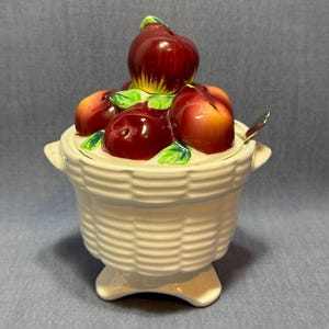 May include: A white ceramic basket-shaped jar with a lid, adorned with a cluster of red and yellow-orange fruit and green leaves. The jar has a small silver spoon and is on a pedestal base.