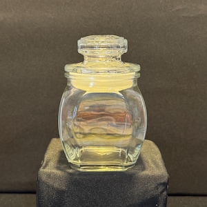 Puede incluir: Un tarro de cristal transparente con base cuadrada y cuerpo redondeado. El tarro tiene una tapa a juego con un diseño decorativo. El tarro está sobre una superficie negra. El tarro probablemente se utiliza para guardar objetos pequeños.