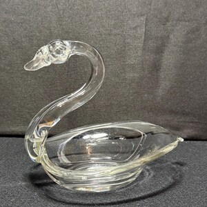 May include: A clear glass swan-shaped dish. The swan has a curved neck and a bowl-shaped body. The dish is made of clear glass and sits on a dark surface. The swan's head is detailed with a beak and eye.