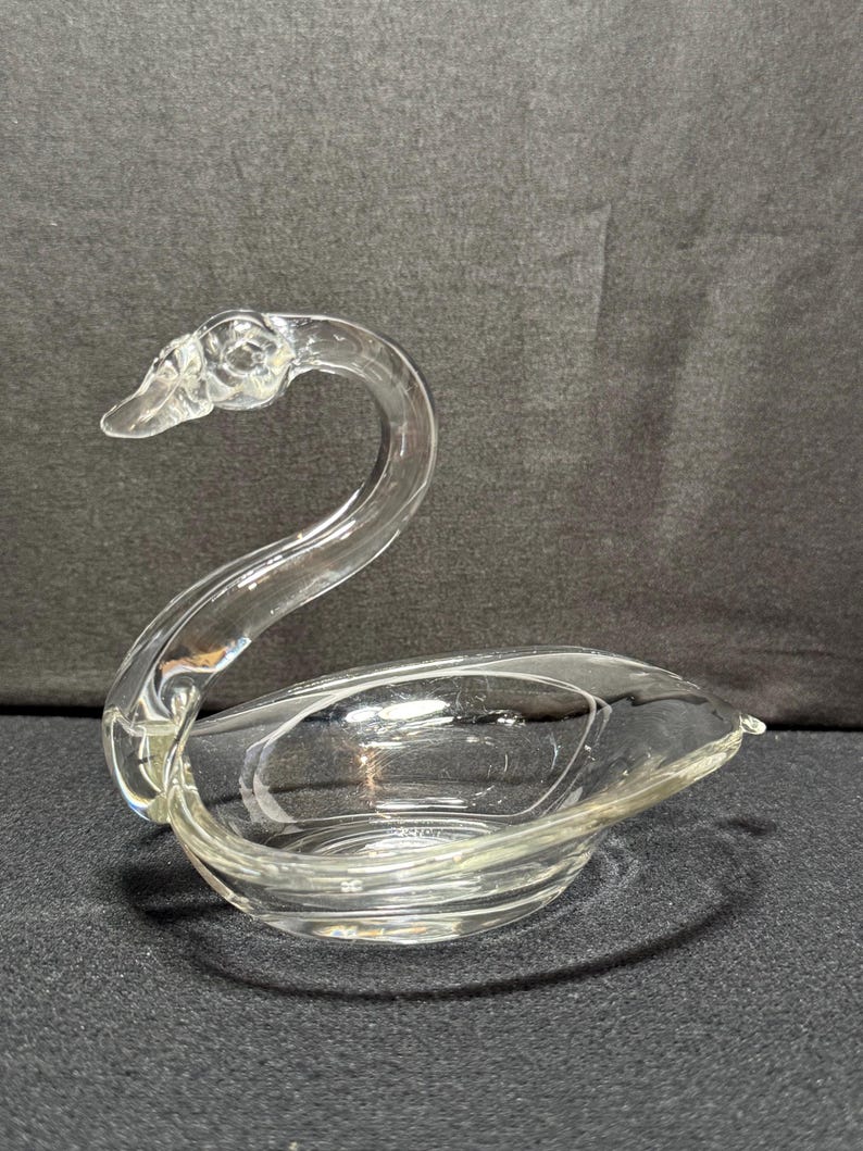 May include: A clear glass swan-shaped dish. The swan has a curved neck and a bowl-shaped body. The dish is made of clear glass and sits on a dark surface. The swan's head is detailed with a beak and eye.