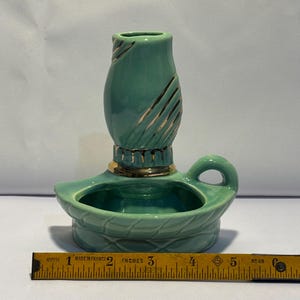 May include: A teal ceramic candle holder with a gold-trimmed base and a handle, featuring a textured pattern on the candle cup. A yellow measuring tape placed in front shows the object is approximately 6 inches wide at its base.