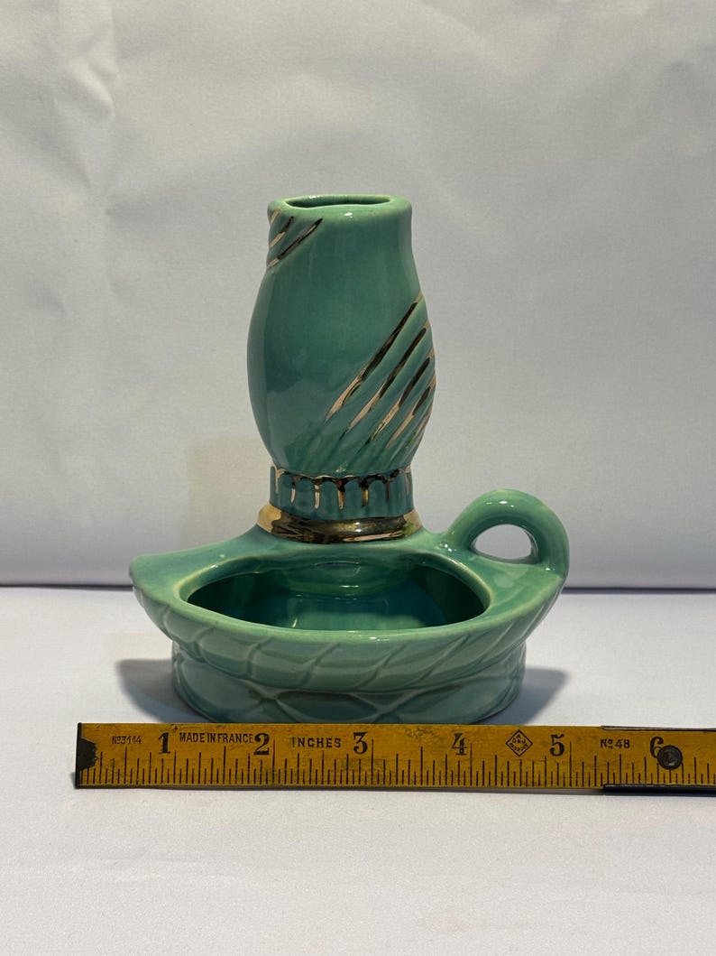 May include: A teal ceramic candle holder with a gold-trimmed base and a handle, featuring a textured pattern on the candle cup. A yellow measuring tape placed in front shows the object is approximately 6 inches wide at its base.