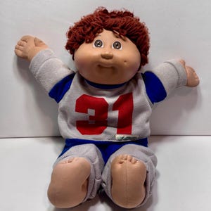 May include: A vintage Cabbage Patch Kid doll, featuring a soft, fabric body and a vinyl head. The doll is wearing a gray and blue football uniform with the number 31 in red. The doll has brown curly hair and open arms.