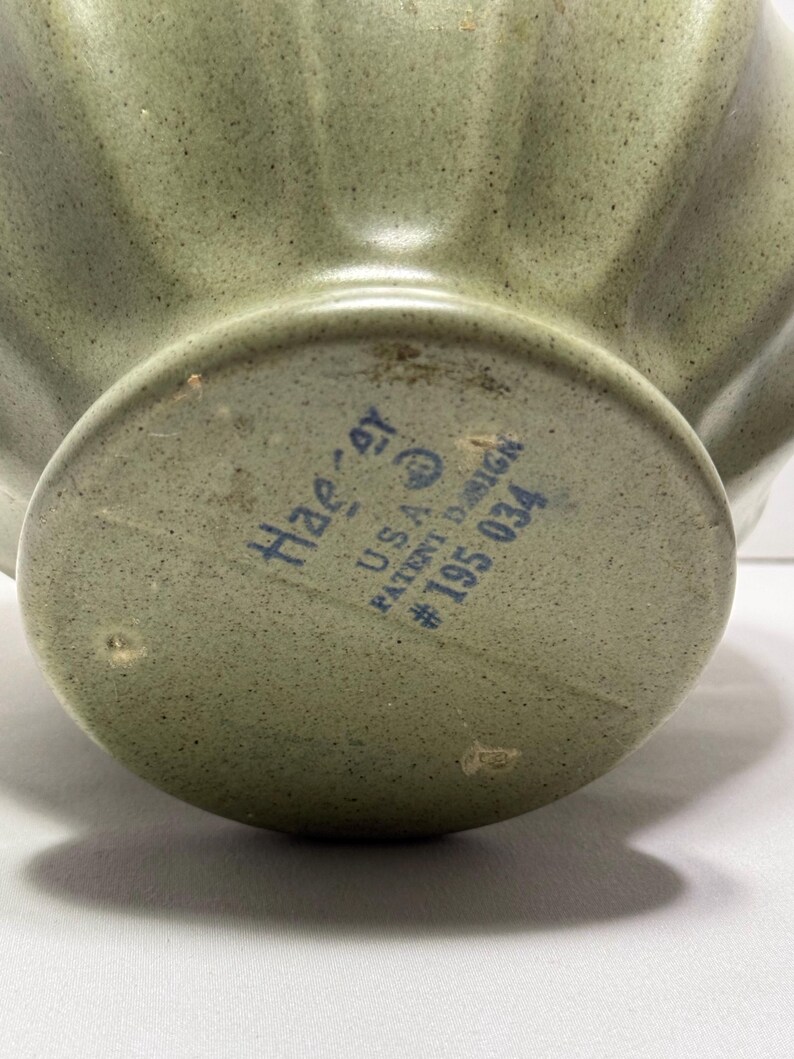 May include: Close-up of a sage green ceramic bowl with a fluted design. The bottom of the bowl has the text "Haeger U.S.A. PATENT DESIGN #195 034" in blue ink. The bowl has a speckled texture.