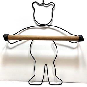 May include: A wall-mounted holder shaped like a bear silhouette, constructed from black wire. The bear figure features a horizontal wooden dowel held across its outstretched arms, designed to support a roll of paper or a towel.