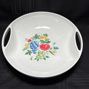 May include: White ceramic serving bowl with two handles. The bowl features a colorful fruit design in the center, including blue grapes, a red apple, and green grapes. The bowl is round and has a glossy finish.