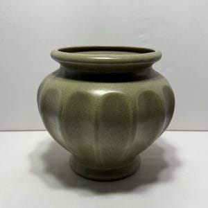 May include: A sage green ceramic vase with a rounded body and a fluted design. The vase has a small, circular base and a slightly flared rim. The overall shape is reminiscent of a pumpkin. The vase is approximately 6 inches tall.