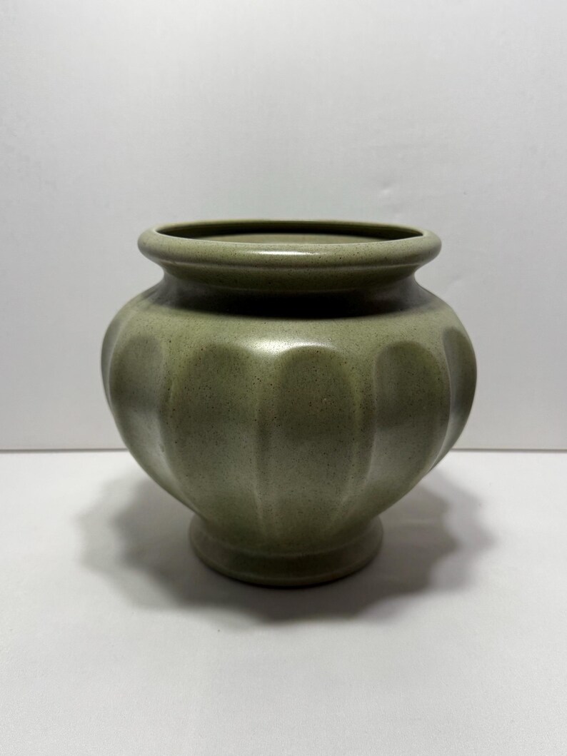 May include: A sage green ceramic vase with a rounded body and a fluted design. The vase has a small, circular base and a slightly flared rim. The overall shape is reminiscent of a pumpkin. The vase is approximately 6 inches tall.