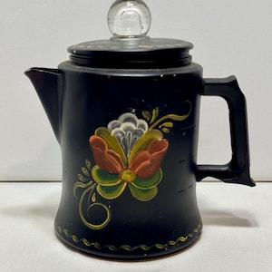 May include: A black coffee percolator featuring a hand-painted floral design in orange, green, and white on the front. The pot includes a black handle, a matching black lid, and a clear glass knob on top. The base is decorated with a wavy line.