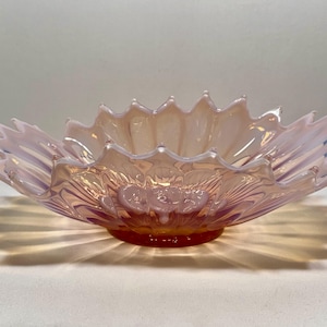 May include: An iridescent glass bowl with a petal-shaped rim. The bowl has a circular base and a light pink colour with orange and purple tones. The design of the bowl creates interesting shadows.