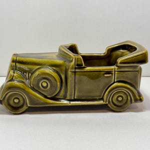 Pode incluir: Um vaso de cerâmica em forma de carro vintage na cor verde-oliva. O vaso apresenta molduras detalhadas de um carro clássico com rodas, faróis e capota conversível visíveis. A parte superior aberta oferece espaço para pequenas plantas ou itens decorativos.