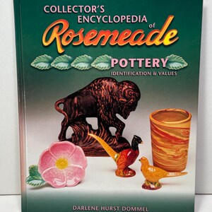 May include: A hardcover book titled Collector's Encyclopedia of Rosemeade Pottery Identification & Values by Darlene Hurst Dommel. The dark green cover features a brown buffalo figurine, a pink flower dish, two bird figurines, and a marbled tumbler.