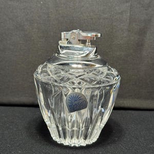 Puede incluir: Un encendedor de mesa de cristal transparente con una parte superior de metal plateado. El encendedor tiene un patrón facetado en forma de diamante en el cuerpo de vidrio. La etiqueta dice "HIGHLIGHTS IN LEAD CRYSTAL BY FRANCESS HOULE".