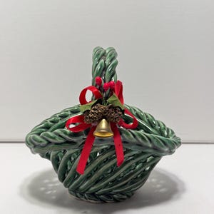 May include: A green ceramic basket with a woven design and a handle. The basket is decorated with a red ribbon, a small gold bell, and pinecones. The basket is a decorative item.