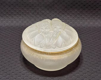 May include: A round, frosted glass trinket box featuring a lid with a molded relief of two birds perched together. The base of the container has a textured, matte finish, and a thin gold-colored metal band encircles the rim where the lid meets the base.