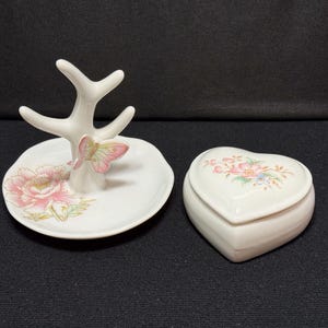 Può includere: Un portagioie in ceramica bianca con un design floreale e un accento a farfalla, accanto a un portagioie a forma di cuore con dettagli floreali. Il supporto ha una struttura ad albero.