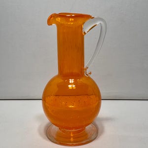 Peut inclure: Une carafe en verre orange vif dotée d'une base bulbeuse avec de petites bulles d'air incrustées, d'un long col cylindrique, d'une anse en verre transparent et d'une base circulaire transparente. La carafe présente un bec verseur distinct.