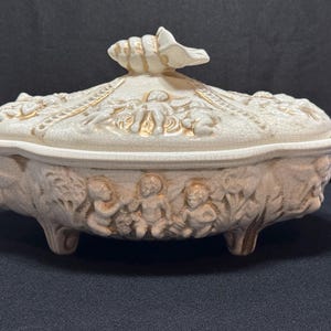 Puede incluir: Joya de cerámica color crema con tapa, con esculturas en relieve de querubines y diseños florales. La caja tiene tres patas pequeñas y un asa en forma de concha. Detalles dorados. Mide aproximadamente 20 cm de largo.