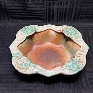 Puede incluir: Una maceta de cerámica con un diseño de borde festoneado único. La maceta presenta un borde verde claro y crema con detalles florales. El interior tiene una combinación de colores marrón y naranja, creando una estética rústica.