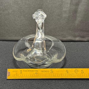 May include: Clear glass swan-shaped dish with a detailed head and neck, and a circular base. The dish is approximately 5 inches long, and is sitting on a dark surface. A yellow ruler is in the foreground.