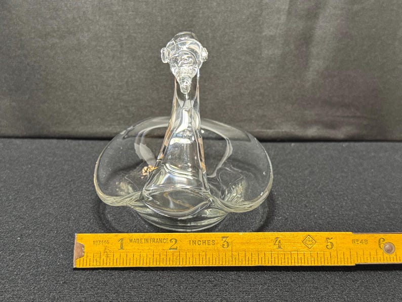 May include: Clear glass swan-shaped dish with a detailed head and neck, and a circular base. The dish is approximately 5 inches long, and is sitting on a dark surface. A yellow ruler is in the foreground.