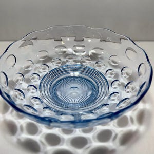 May include: A light blue glass bowl with a scalloped edge and a pattern of circular indentations. The center of the bowl has a textured, circular design. The bowl is sitting on a white surface.