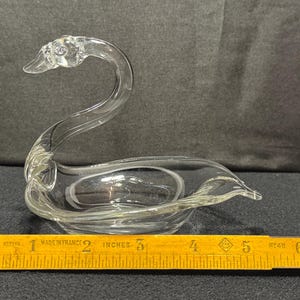 May include: A clear glass swan figurine with a curved neck and a bowl-shaped body. The swan is approximately 6 inches long, measured with a wooden ruler. The figurine is likely a decorative item.