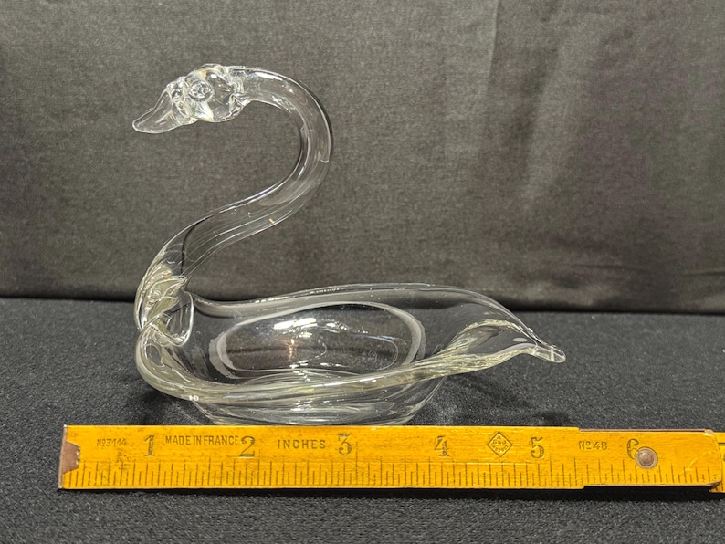 May include: A clear glass swan figurine with a curved neck and a bowl-shaped body. The swan is approximately 6 inches long, measured with a wooden ruler. The figurine is likely a decorative item.