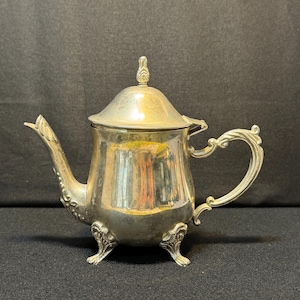 Puede incluir: Tetera plateada con tapa y asa ornamentada. La tetera tiene un pico curvo y se apoya en tres patas decorativas. El diseño es vintage, con un estilo clásico y elegante. La tetera es de metal y refleja la luz.