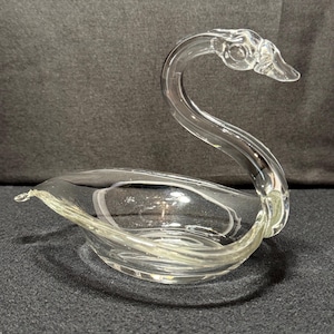 May include: A clear glass swan-shaped dish. The swan has a curved neck and a bowl-shaped body. The glass is transparent, allowing light to pass through. The dish is designed to hold small items or serve as a decorative piece.