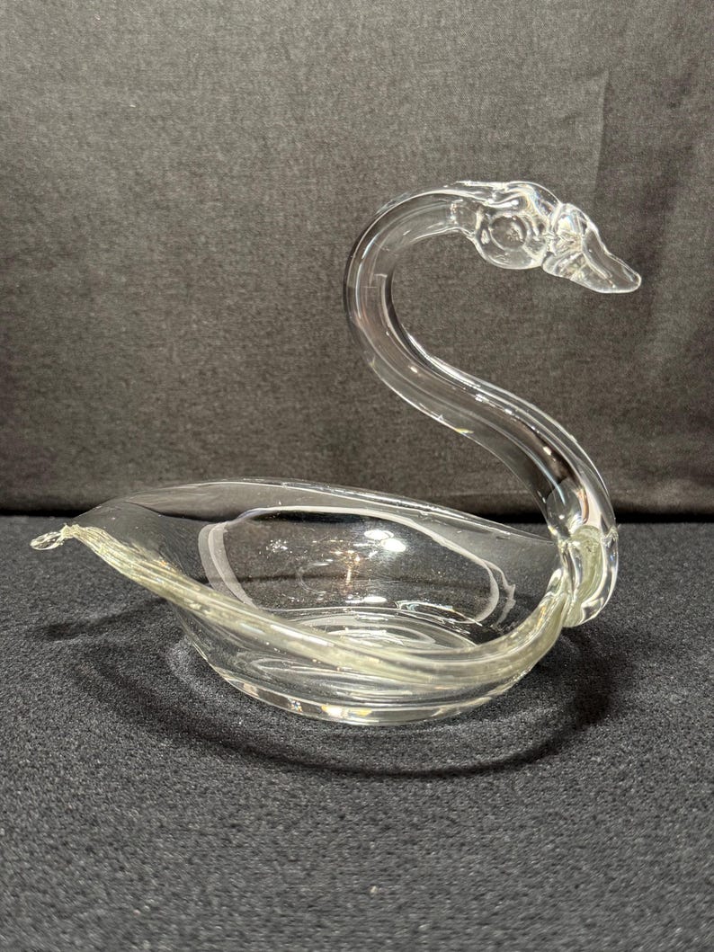 May include: A clear glass swan-shaped dish. The swan has a curved neck and a bowl-shaped body. The glass is transparent, allowing light to pass through. The dish is designed to hold small items or serve as a decorative piece.