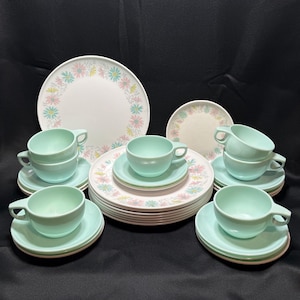 May include: A dinnerware set featuring white plates with a floral pattern of pink, blue, and yellow daisies. The set includes several solid light blue cups and matching saucers, along with a stack of patterned plates and a single small plate.