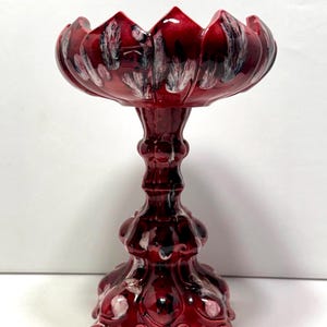 May include: A pedestal bowl featuring a deep red glaze with white and black mottled accents. The bowl has a scalloped rim and sits on a decorative, multi-tiered base with ornate, curved details. The ceramic surface has a glossy finish.