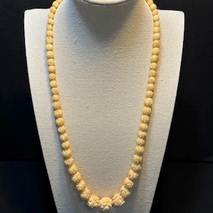 May include: Cream-colored beaded necklace displayed on a mannequin. The necklace features small, round beads and larger, intricately carved floral-shaped beads. The beads are strung together, creating a delicate and elegant piece of jewelry.