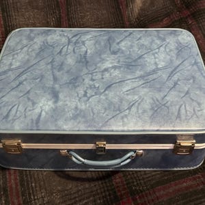 May include: A rectangular blue suitcase with a marbled texture, featuring a white trim along the center, a light blue handle, and three silver-toned metal latches on the front. The suitcase has a structured, hard-shell design.