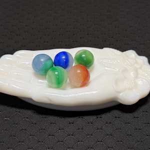 Op de afbeelding: Een witte melkglazen schaal in de vorm van een open hand met bloemmotieven bij de pols, met daarin vijf kleine, bolvormige glazen knikkers in blauwe, groene en oranje tinten. De schaal heeft een gladde, ondoorzichtige afwerking en een gebogen vorm.
