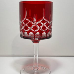 May include: A red crystal wine glass with a clear stem and base. The glass features a geometric pattern cut into the red glass. The stem is clear, and the base is round. The glass is designed for serving wine.