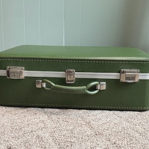 May include: A rectangular olive green suitcase featuring a smooth exterior finish, a central handle, and three silver-toned metal latches along the front edge. The suitcase is constructed with a reinforced frame and visible stitching details.